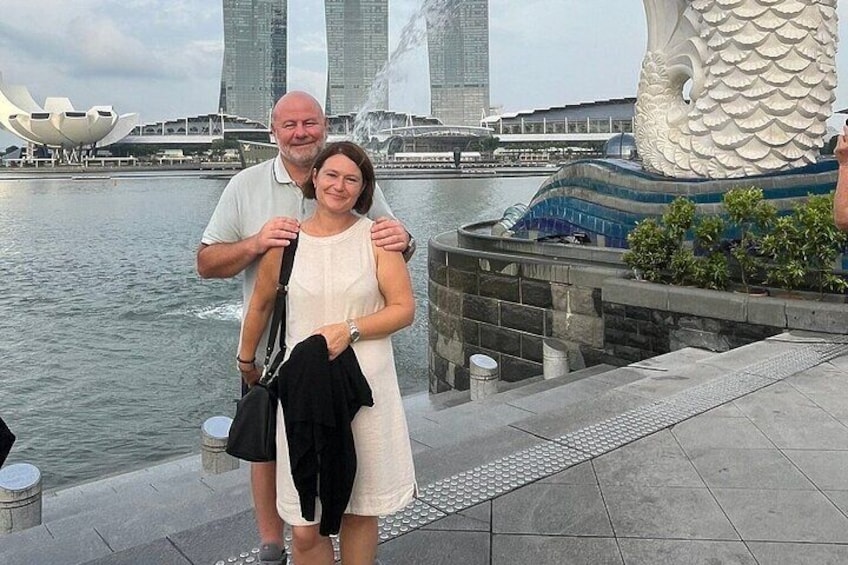 A couple standing by a waterfront with a large dragon sculpture and modern buildings in the background. 4