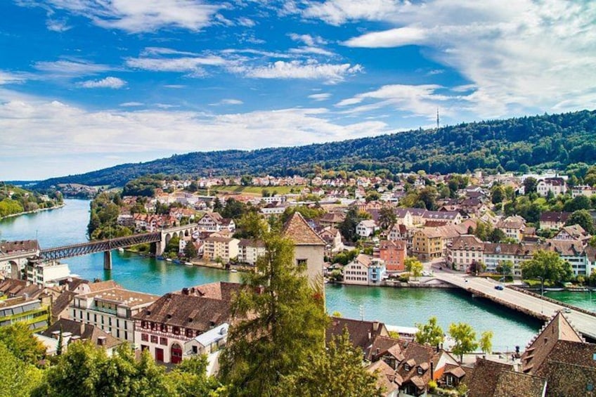 A riverside town with a bridge, buildings, and a hill in the background. 1