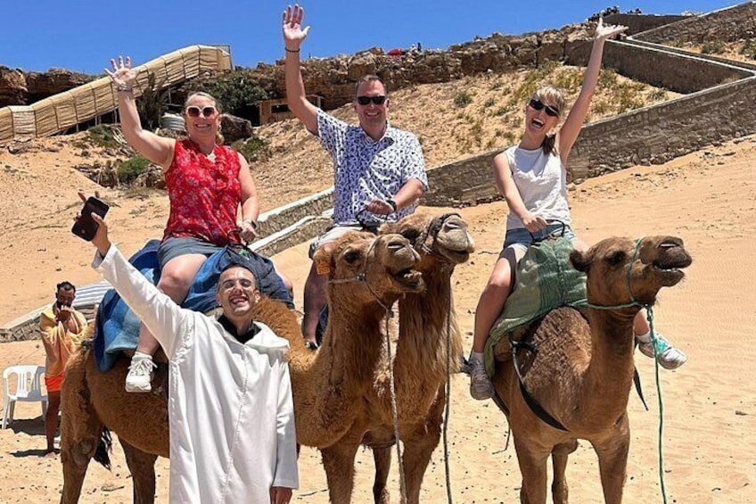 Personas montando camellos en un paisaje desértico, con una estructura de madera al fondo. 5