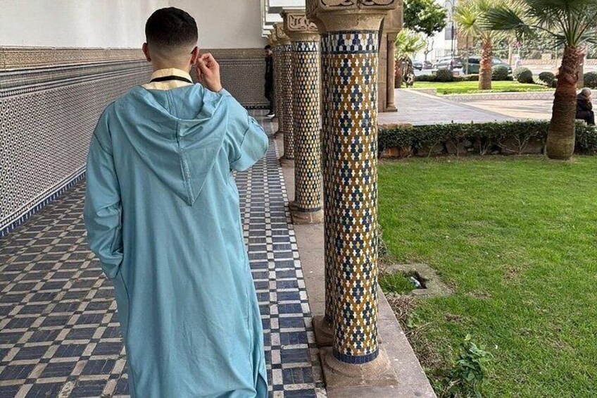 Una persona con una bata azul caminando sobre un suelo de baldosas ornamentadas, con columnas decorativas y un área verde al fondo. 4