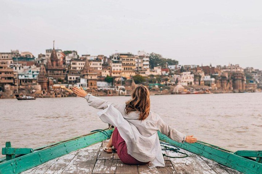 A person sitting on a wooden dock with arms outstretched, overlooking a river with buildings in the background. 1
