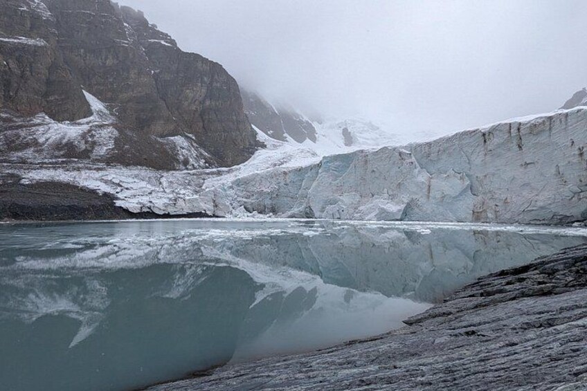 A glacier with a reflective lake, rocky cliffs, and snow-covered mountains. 2
