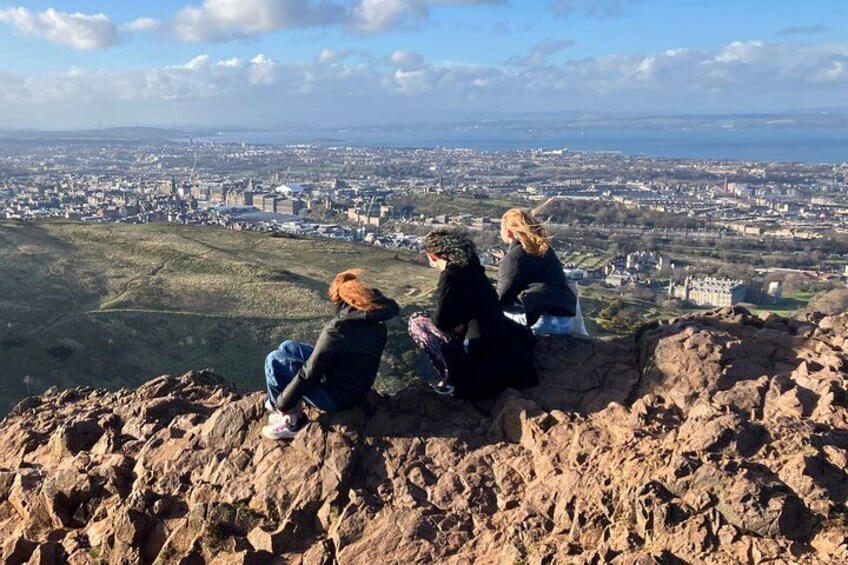 Three people sitting on a rocky outcrop overlooking a cityscape. 1