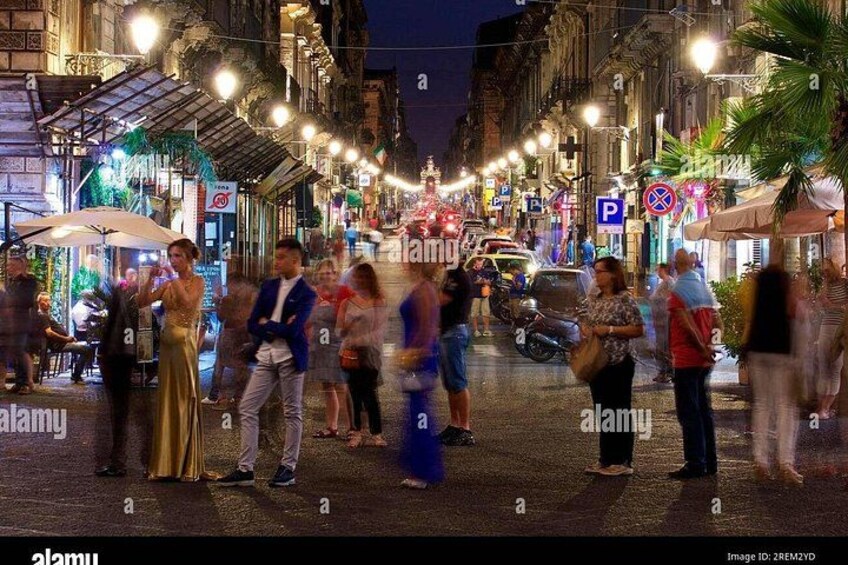 Una calle concurrida de noche, con gente caminando, coches estacionados y carteles iluminados. 4