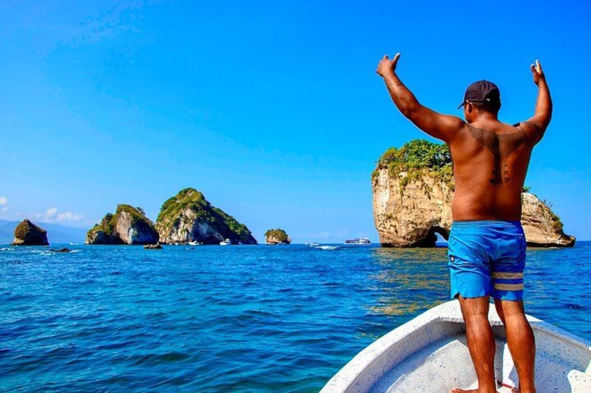 Un homme se tient sur un bateau, les bras levés, face à une mer bleue et dégagée, avec des îles rocheuses en arrière-plan. 5