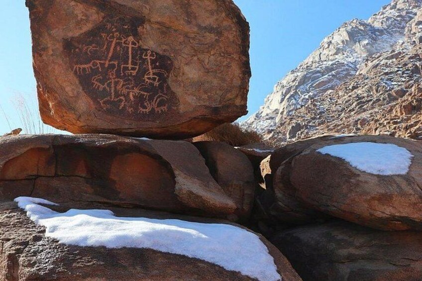 A large rock with ancient petroglyphs, snow on nearby rocks, and a rugged mountainous backdrop. 3