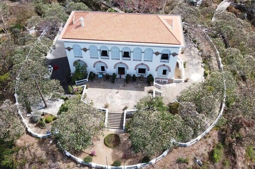 An aerial view of a white building with a tiled roof, surrounded by trees and a paved area. 3