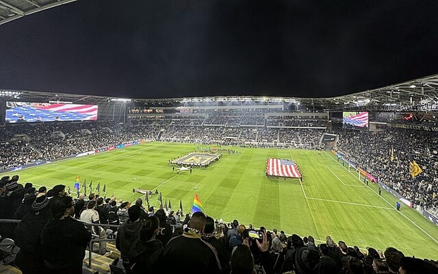 A stadium filled with spectators, a large American flag, and a rainbow flag on the field. 1