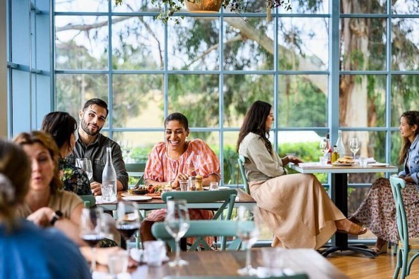 En gruppe mennesker spiser på en udendørs restaurant med store vinduer og hængende planter. 2