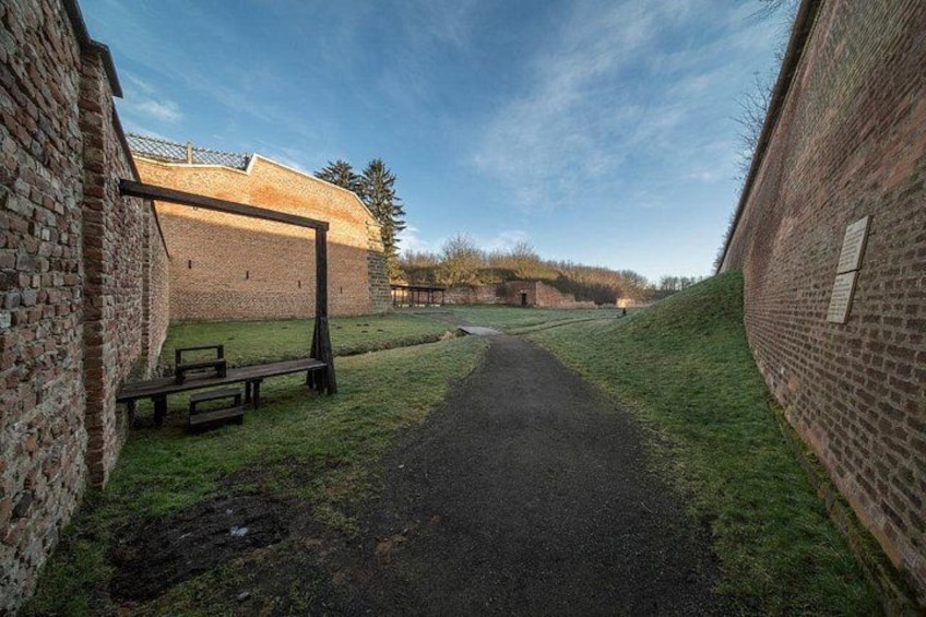 A pathway leading through a brick wall, with a grassy area and a bench on the left. 2