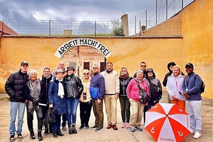 A group of people standing in front of a building with a sign that reads 'ARBEIT MACHT FREI'. 1