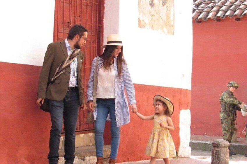 A family with a child holding hands, standing in front of a red and white building with a soldier in the background. 3