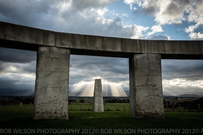A large concrete structure with three pillars and a central column, set against a cloudy sky with sunlight breaking through. 2