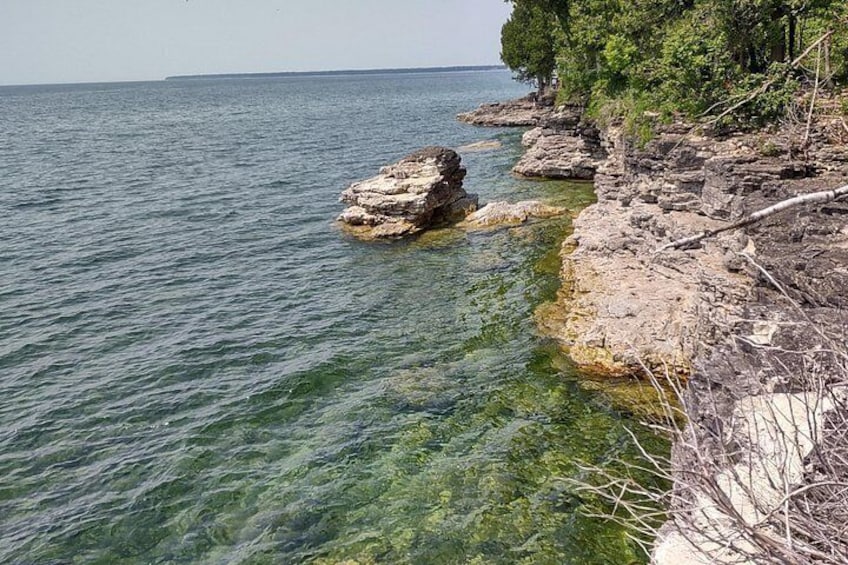 A rocky shoreline with clear water, a distant tree line, and a clear sky. 3