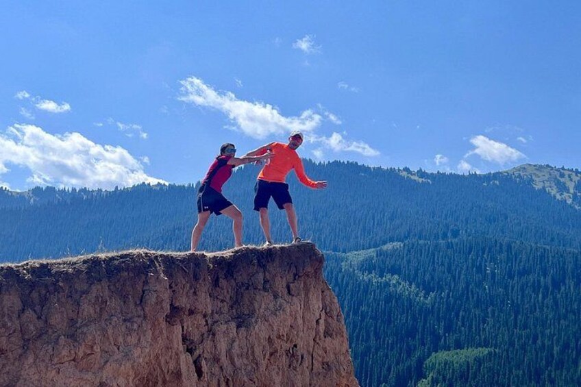 Two people standing on a cliff edge with a forested mountain range in the background. 2
