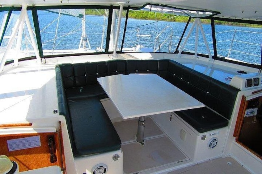 A boat's interior with a cushioned seating area, a table, and a view of the water through the windows. 2