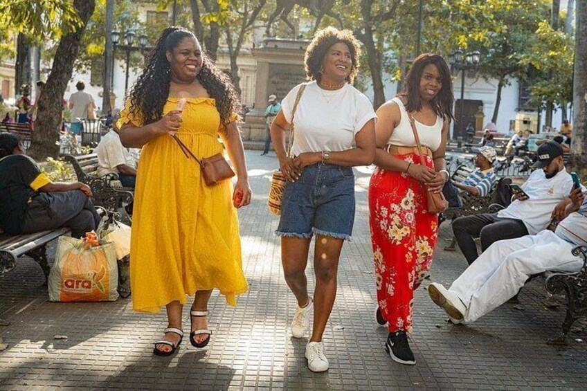 Tre donne che camminano su un vialetto lastricato in un parco, sullo sfondo alberi e panchine. 1