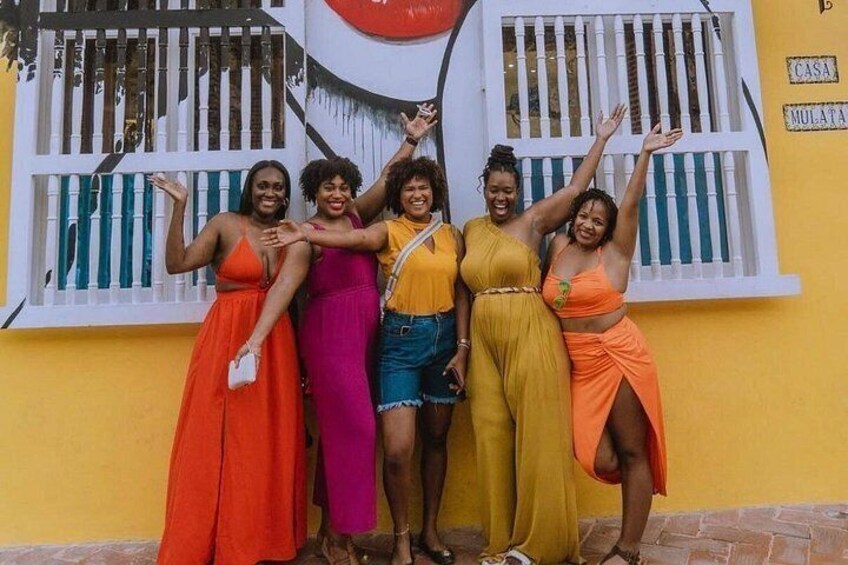 Un gruppo di donne con abiti colorati in piedi di fronte a un edificio con un murale e una finestra con sbarre. 5