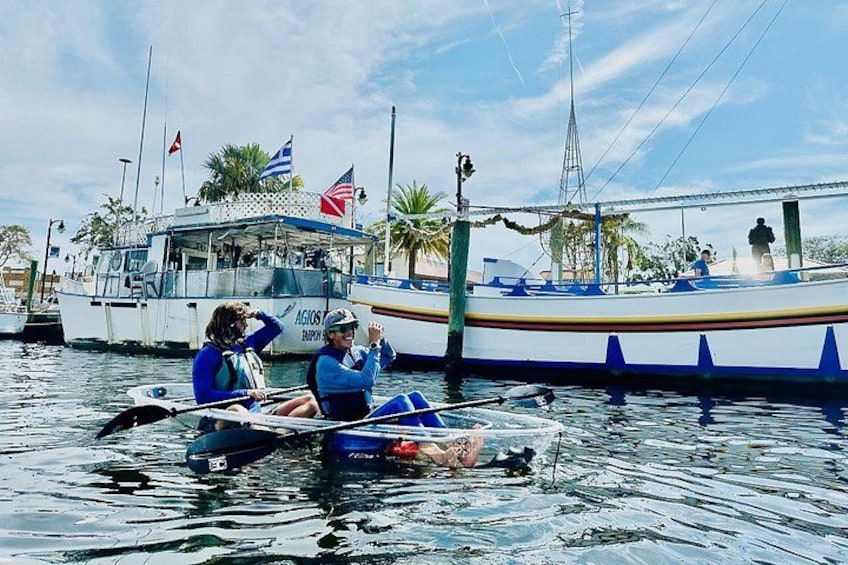 Two people in a small boat on calm water, a larger boat with flags in the background, and a dock with palm trees. 1
