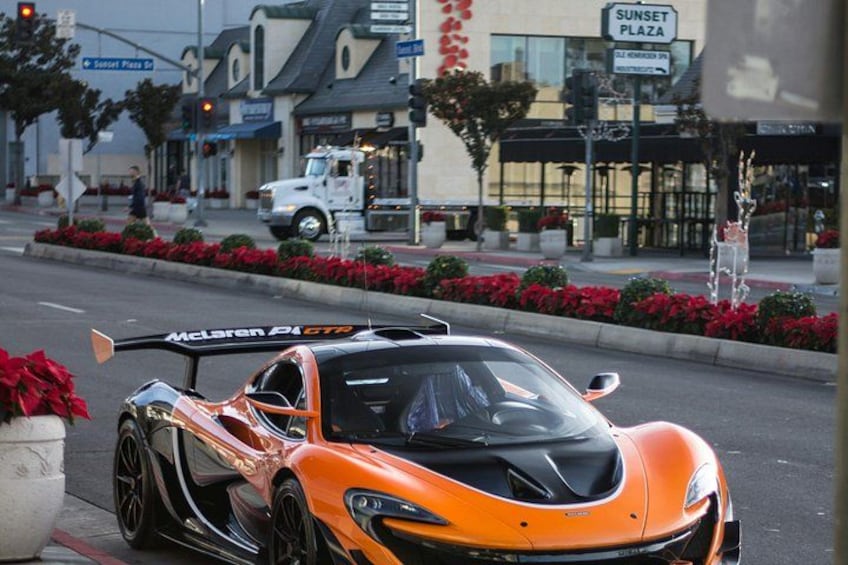Een oranje sportauto geparkeerd langs de kant van een straat, met een gebouw genaamd 'Sunset Plaza' op de achtergrond. 2
