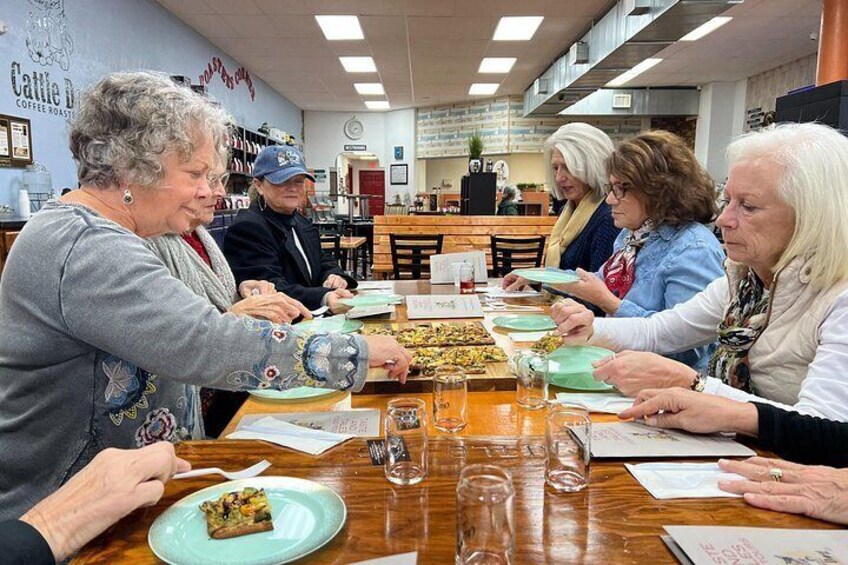 A group of people sitting around a table with plates of food, in a cafe with a coffee roasting sign on the wall. 2