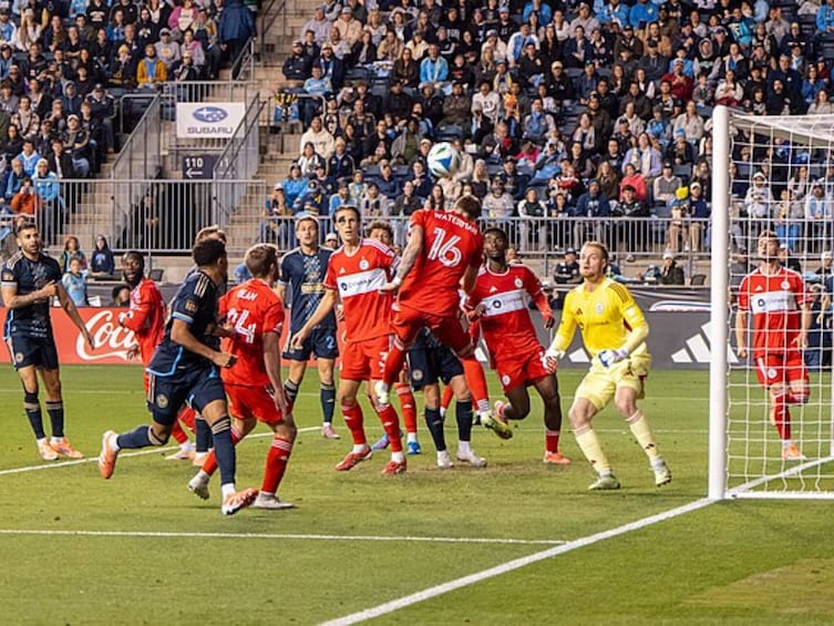 Ein Fußballspiel mit Spielern in roten und blauen Trikots, einem gelben Torwart und einer Zuschauermenge im Hintergrund. 3