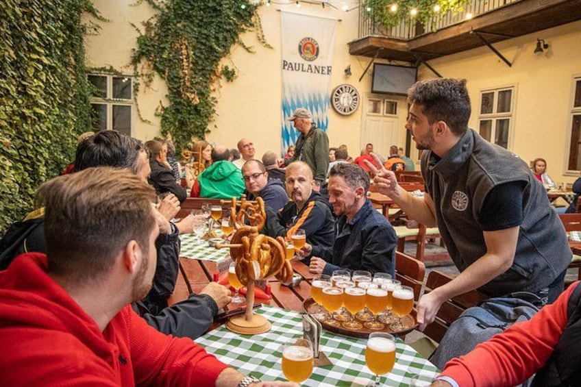 A group of people sitting at a table with beer glasses, surrounded by greenery and a banner. 2