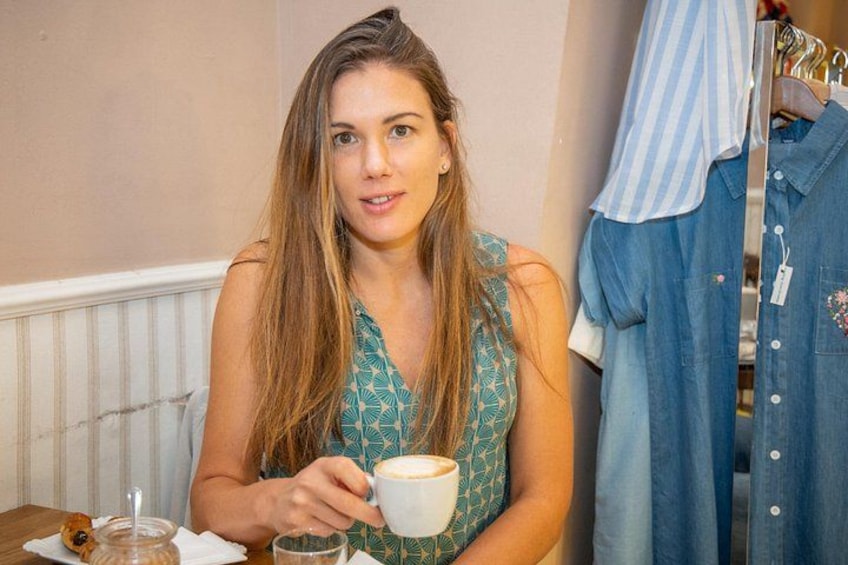 A person sitting at a table with a cup of coffee, a pastry, and a glass of water. There is a clothing rack with hanging garments in the background. 5