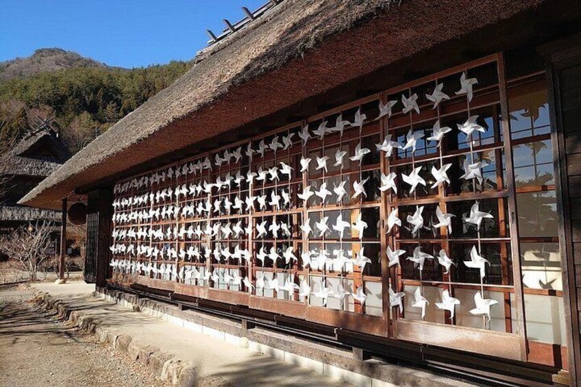 Ein traditionelles japanisches Gebäude mit einem strohgedeckten Dach und einer Wand, die mit zahlreichen weißen Papierkranichen verziert ist. 4