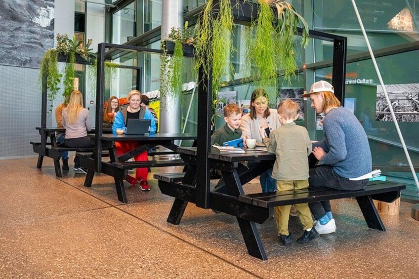 People sitting at tables with laptops and books, in a modern indoor setting with glass walls and hanging plants. 3