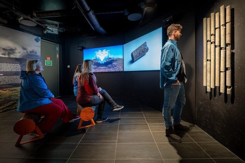 A museum exhibit with a large screen displaying a landscape, a wall with cylindrical objects, and visitors seated and standing. 4