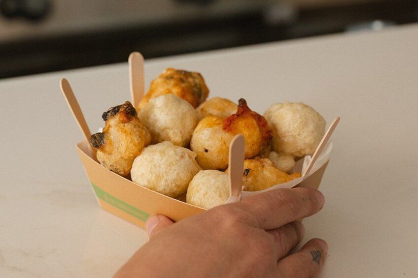 Una mano che tiene un contenitore di carta con fritture rotonde e dorate. 5