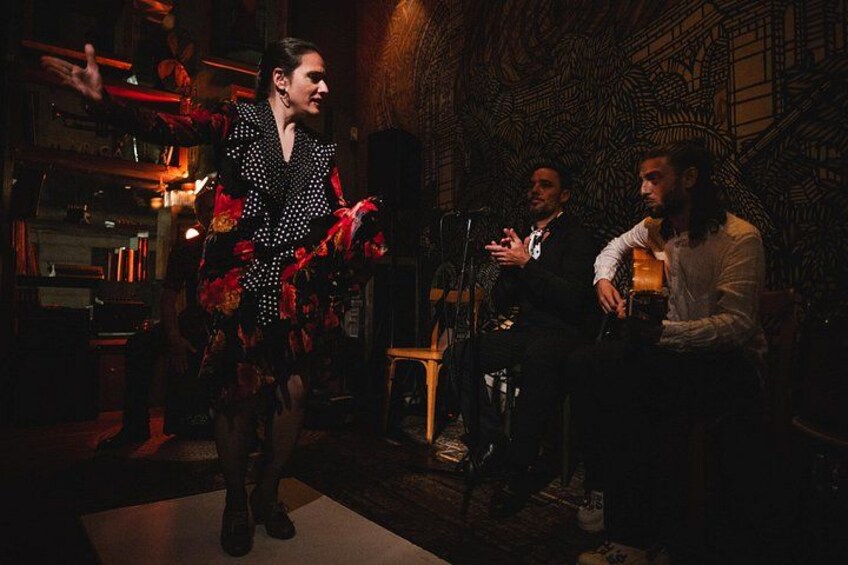 Una persona con un vestido rojo y negro estampado, un hombre con traje y otro con camisa blanca en una habitación poco iluminada con papel tapiz estampado. 2