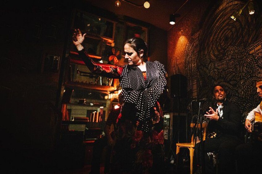 A person performing on stage with a microphone, a patterned wall, and a bookshelf in the background. 2
