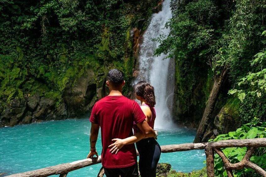 Una pareja de pie junto a una cascada turquesa rodeada de exuberante vegetación. 1