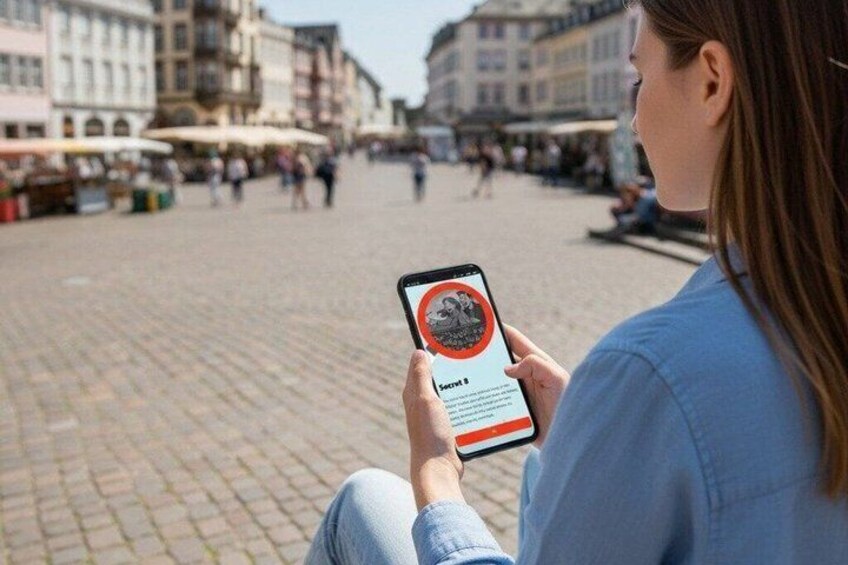 A person sitting on a cobblestone square, holding a smartphone with a red circle and text on the screen. 5