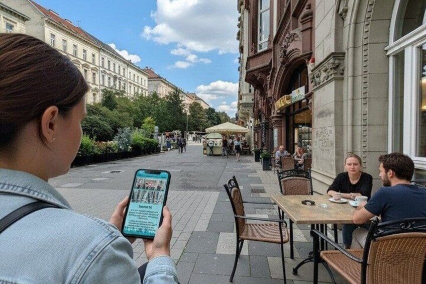 A person using a smartphone on a city street with buildings and outdoor seating. 1
