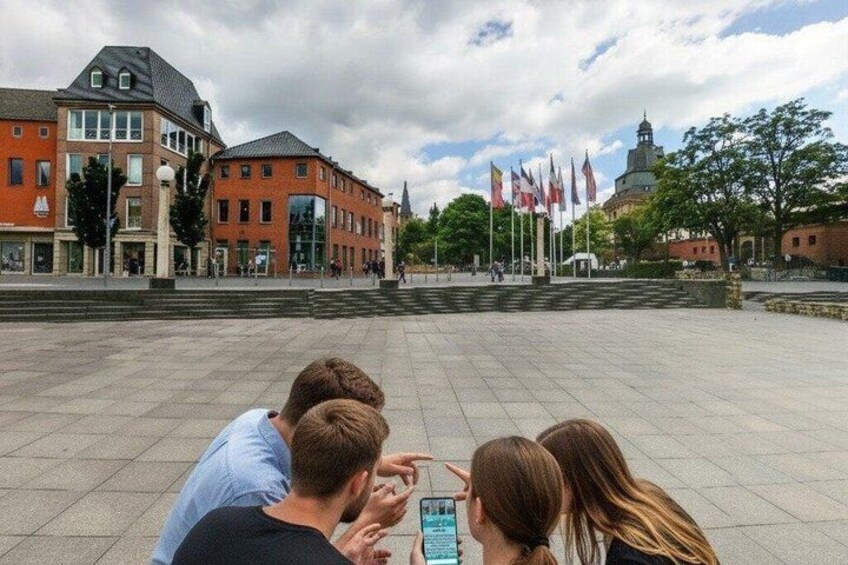 A group of people looking at a smartphone in a public square with buildings and flags in the background. 4