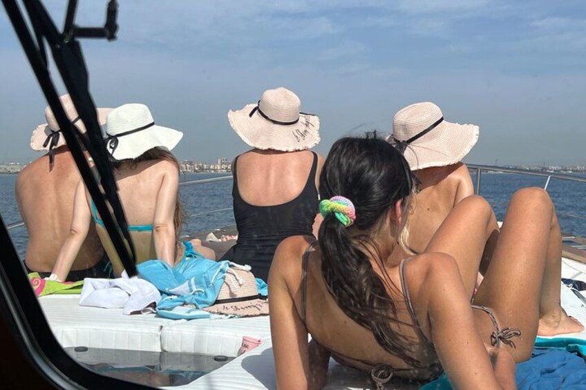 People on a boat, some wearing hats, with a body of water and a distant shoreline in the background. 2