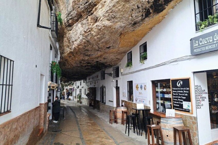A narrow street with a rock overhang, a café with outdoor seating, and a signboard with text. 4