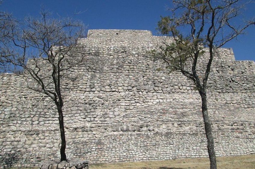 Una gran estructura de piedra con una parte superior plana y una base hecha de piedras más pequeñas. Dos árboles se encuentran frente a la estructura. 3