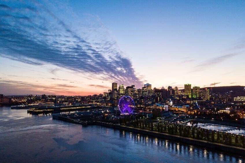 A city skyline at dusk with illuminated buildings, a prominent Ferris wheel, and a body of water reflecting the lights. 2