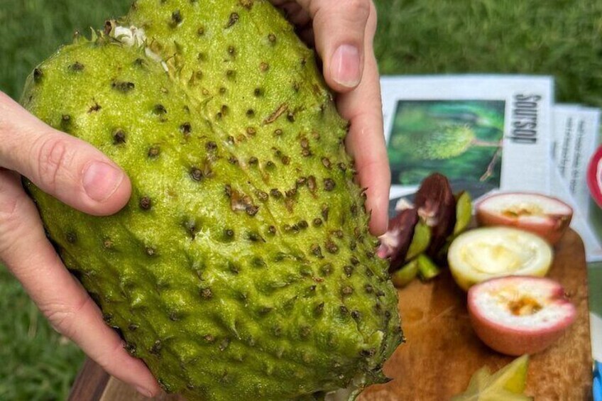 A person holding a green, bumpy fruit with a spiky texture, and a wooden cutting board with sliced fruit in the background. 5