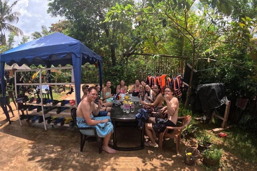 Een groep mensen zit buiten aan een tafel, met een blauwe tent en diverse spullen die aan een rek hangen. 4