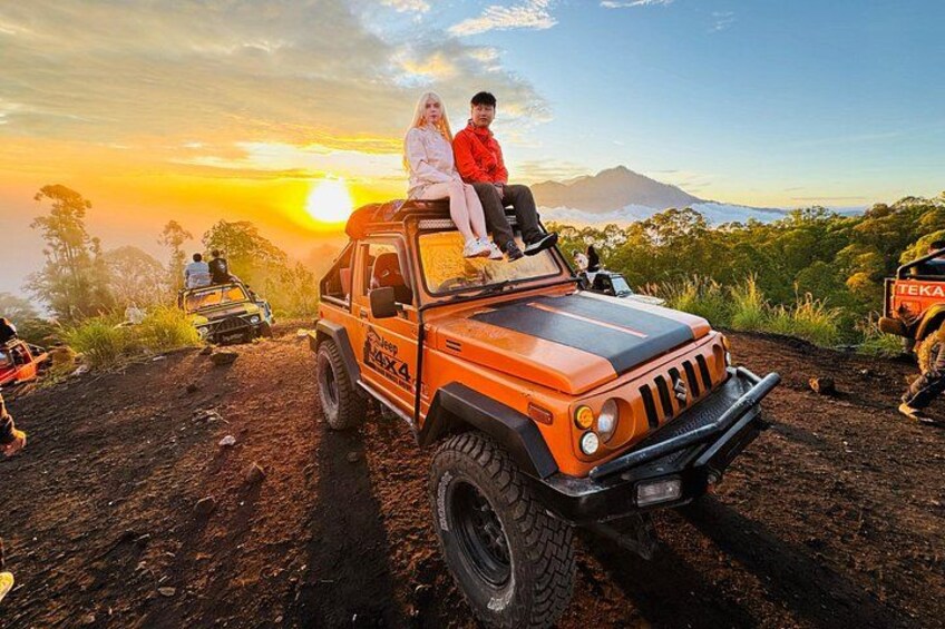 A jeep with two passengers on a dirt road, a sunset in the background, and a mountain. 1