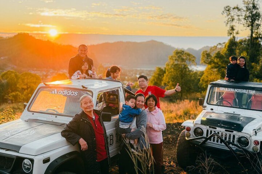 A group of people around two white SUVs with 'adidas' branding, parked on a hill with a sunset in the background. 2