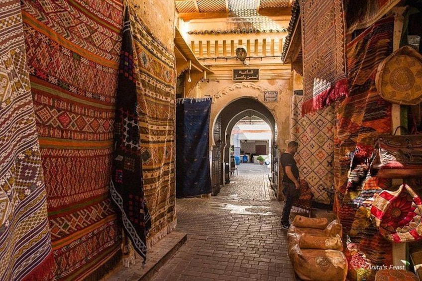 Un callejón estrecho con alfombras y textiles de diseños variados expuestos, un arco y una persona parada a lo lejos. 4