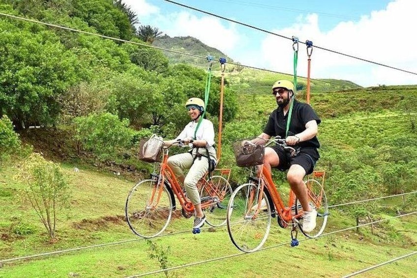 Two people on bicycles suspended in the air by cables, with a green landscape in the background. 1