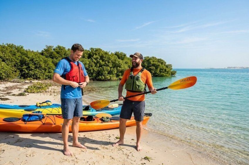 Zwei Personen stehen an einem sandigen Strand mit Kajaks und Stand-Up-Paddles, einem ruhigen Meer und einem klaren Himmel. 2