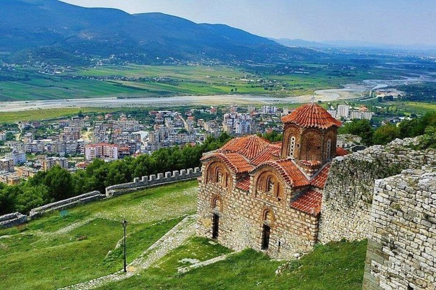 A historic church with red-tiled roofs, a stone wall, and a view of a cityscape with buildings and mountains in the background. 4